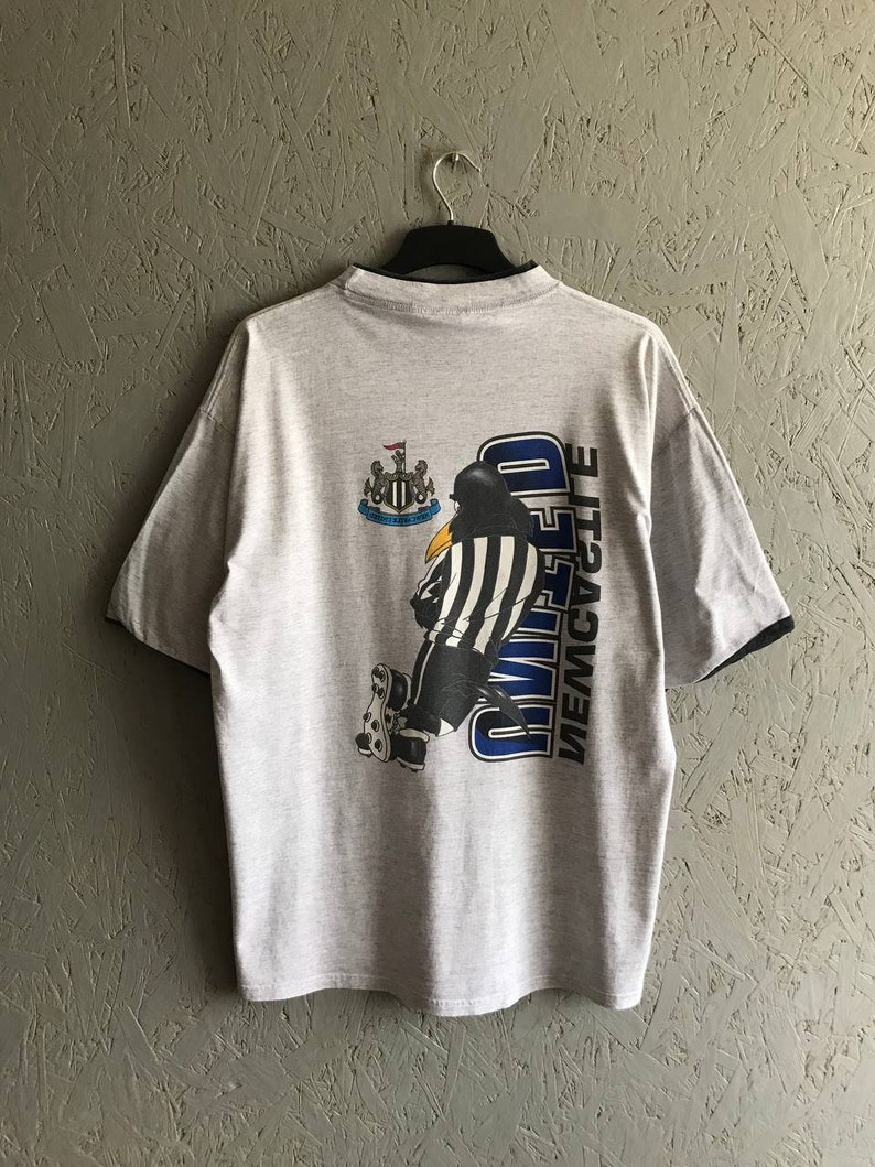 Puede incluir: Una camiseta gris con un dibujo animado en blanco y negro de un ping&uuml;ino patinando con un uniforme de &aacute;rbitro. El texto "NEWCASTLE UNITED" est&aacute; impreso verticalmente en la camiseta.