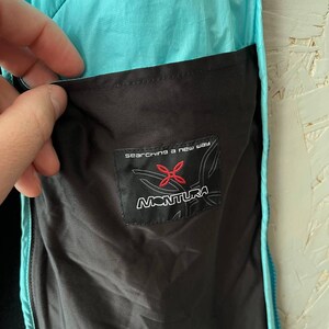 May include: Close-up of a turquoise and black jacket pocket. The pocket features a black label with the brand name "MONTURA" and the text "searching a new way". The jacket is made of a lightweight, water-resistant material.