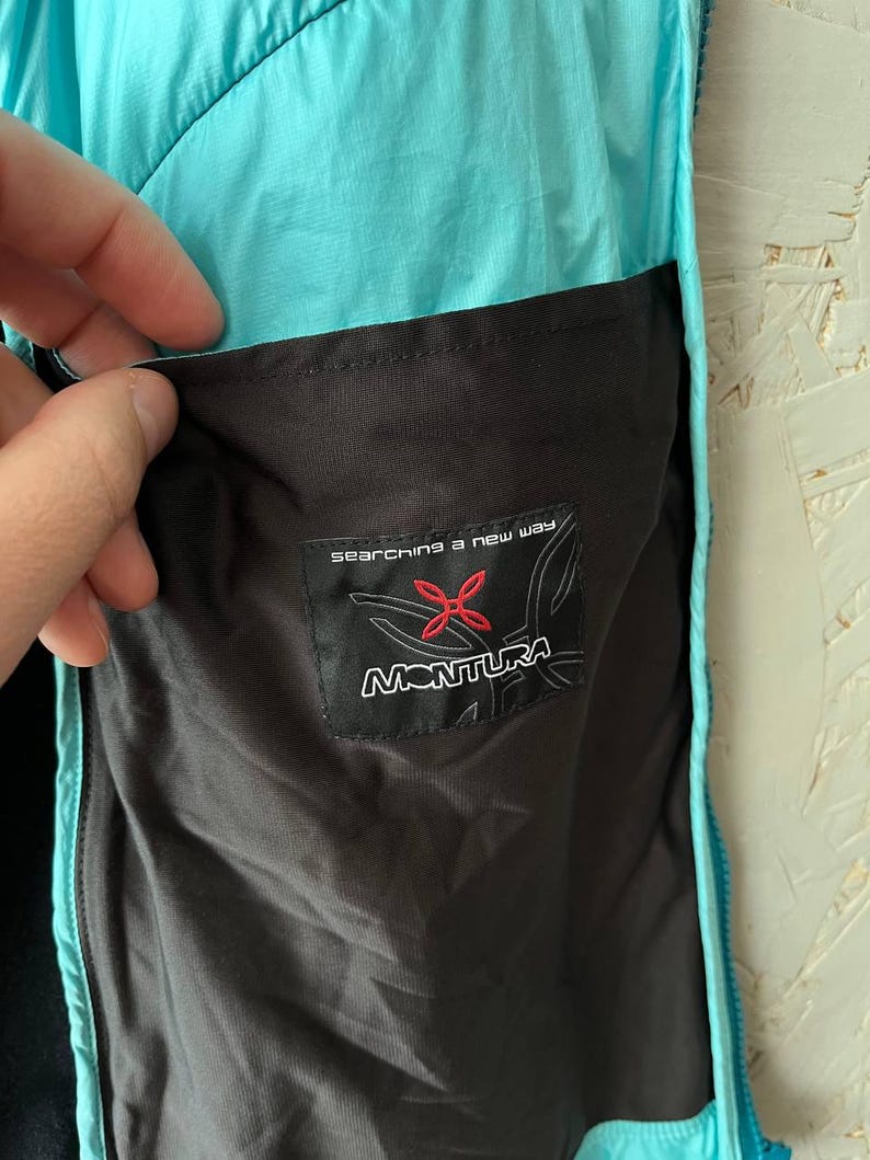 May include: Close-up of a turquoise and black jacket pocket. The pocket features a black label with the brand name "MONTURA" and the text "searching a new way". The jacket is made of a lightweight, water-resistant material.