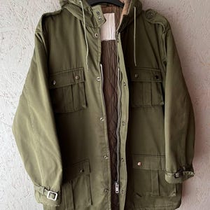 K&ouml;nnte beinhalten: Eine olivgr&uuml;ne Funktionsjacke mit gestepptem braunem Innenfutter, einer Kapuze mit Kordelzug sowie einem Frontrei&szlig;verschluss mit Druckkn&ouml;pfen. Die Jacke verf&uuml;gt &uuml;ber vier gro&szlig;e Vordertaschen, Schulterklappen und verstellbare &Auml;rmelb&uuml;ndchen mit Schnallen.