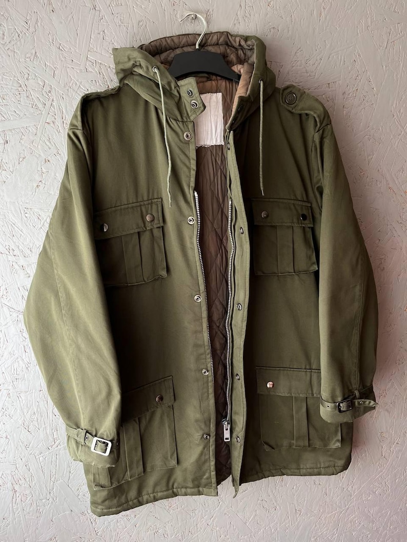 K&ouml;nnte beinhalten: Eine olivgr&uuml;ne Funktionsjacke mit gestepptem braunem Innenfutter, einer Kapuze mit Kordelzug sowie einem Frontrei&szlig;verschluss mit Druckkn&ouml;pfen. Die Jacke verf&uuml;gt &uuml;ber vier gro&szlig;e Vordertaschen, Schulterklappen und verstellbare &Auml;rmelb&uuml;ndchen mit Schnallen.