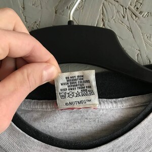 Puede incluir: Una camiseta gris con cuello negro y una etiqueta de cuidado blanca que dice "No planchar la decoraci&oacute;n, Lavar los colores oscuros por separado, Mantener alejado del fuego" y "@NUTMEGTm".