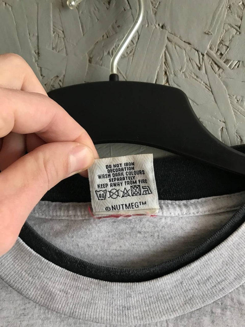 Puede incluir: Una camiseta gris con cuello negro y una etiqueta de cuidado blanca que dice "No planchar la decoraci&oacute;n, Lavar los colores oscuros por separado, Mantener alejado del fuego" y "@NUTMEGTm".