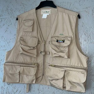 May include: A tan utility vest featuring multiple pockets, a front zipper closure, and a collarless neckline. The L.L.Bean brand label is at the neck, with a small embroidered name JESS and a colorful L.L.Bean logo patch on the upper left pocket.