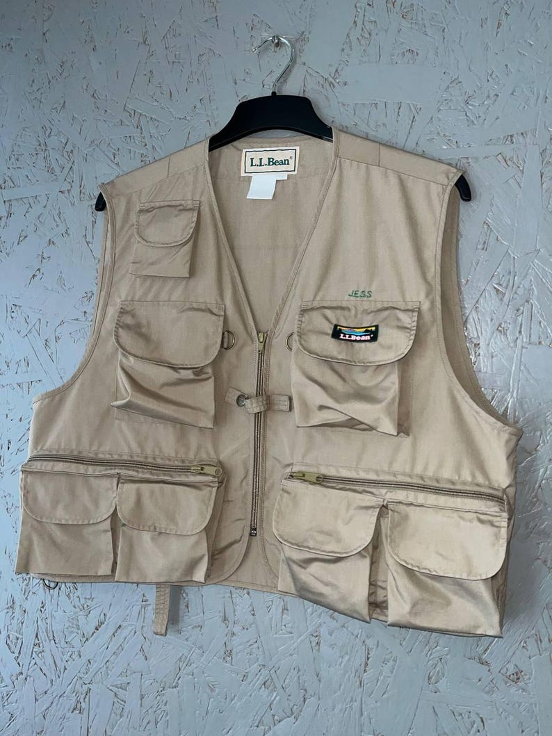 May include: A tan utility vest featuring multiple pockets, a front zipper closure, and a collarless neckline. The L.L.Bean brand label is at the neck, with a small embroidered name JESS and a colorful L.L.Bean logo patch on the upper left pocket.