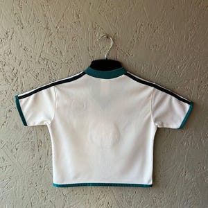 K&ouml;nnte beinhalten: Wei&szlig;es Sport-T-Shirt mit schwarzen Streifen auf den Schultern und t&uuml;rkisfarbenen Verzierungen. Das Shirt hat einen Stehkragen und kurze &Auml;rmel. Es h&auml;ngt an einem Metallb&uuml;gel vor einem strukturierten grauen Hintergrund.