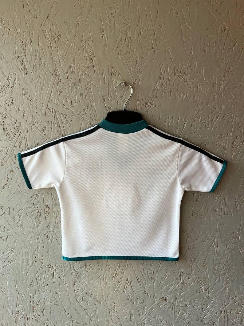 K&ouml;nnte beinhalten: Wei&szlig;es Sport-T-Shirt mit schwarzen Streifen auf den Schultern und t&uuml;rkisfarbenen Verzierungen. Das Shirt hat einen Stehkragen und kurze &Auml;rmel. Es h&auml;ngt an einem Metallb&uuml;gel vor einem strukturierten grauen Hintergrund.