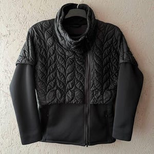 May include: A black quilted jacket with a high collar and a full-length zipper. The jacket features a unique leaf-like quilted pattern on the upper portion and solid black sleeves and lower section. It is hanging on a black hanger.