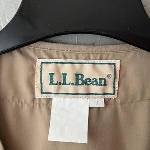 May include: A tan-colored garment featuring a rectangular white fabric label with a green border and the text L.L.Bean in green lettering. The label is stitched onto the interior neckline of the garment, which is suspended from a black hanger.