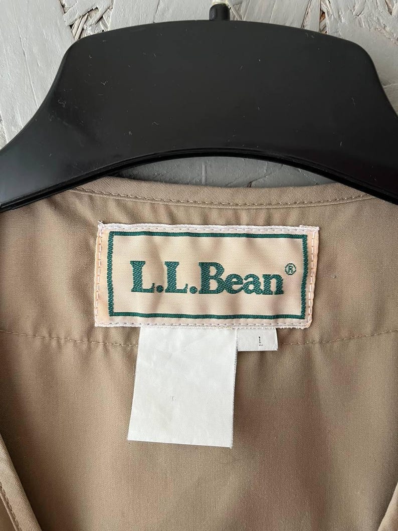 May include: A tan-colored garment featuring a rectangular white fabric label with a green border and the text L.L.Bean in green lettering. The label is stitched onto the interior neckline of the garment, which is suspended from a black hanger.