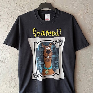 Puede incluir: Camiseta negra con una imagen enmarcada de Scooby-Doo y la palabra "framed!" en amarillo. El perro de dibujos animados lleva un collar azul y una etiqueta. La camiseta está colgada en una percha negra.