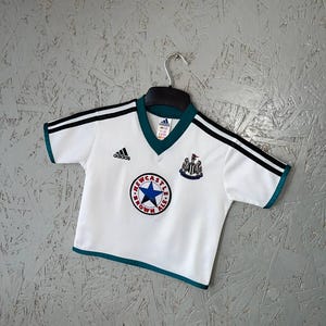K&ouml;nnte beinhalten: Wei&szlig;es Fu&szlig;balltrikot mit schwarzen Streifen auf den &Auml;rmeln und gr&uuml;nen Verzierungen. Das Trikot zeigt das Adidas-Logo und ein rundes Emblem mit einem blauen Stern und der Aufschrift "Newcastle Brown Ale".