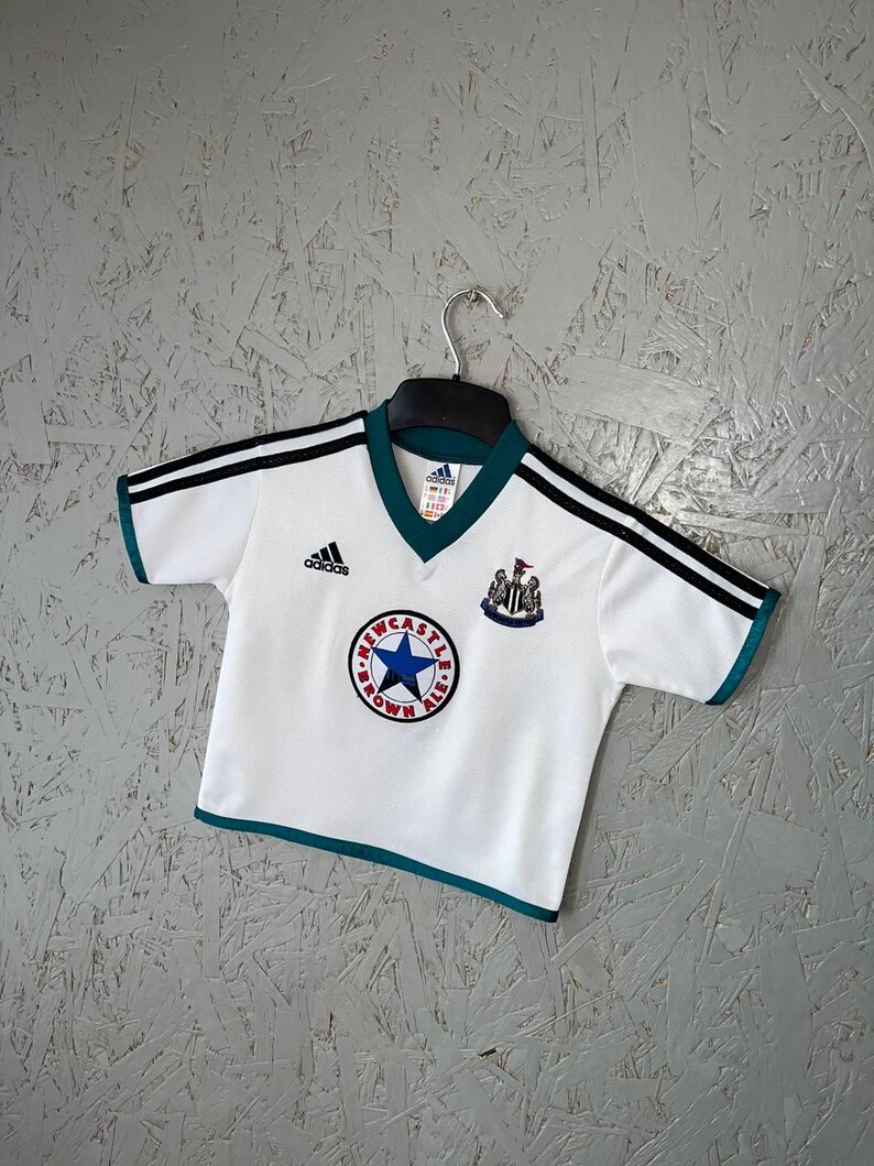 K&ouml;nnte beinhalten: Wei&szlig;es Fu&szlig;balltrikot mit schwarzen Streifen auf den &Auml;rmeln und gr&uuml;nen Verzierungen. Das Trikot zeigt das Adidas-Logo und ein rundes Emblem mit einem blauen Stern und der Aufschrift "Newcastle Brown Ale".