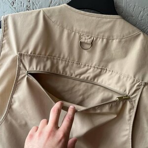 May include: A beige utility vest featuring a sleeveless design with a round neckline. The back of the garment includes a large horizontal zippered pocket and a metal D-ring attachment located just below the collar area for added functionality.