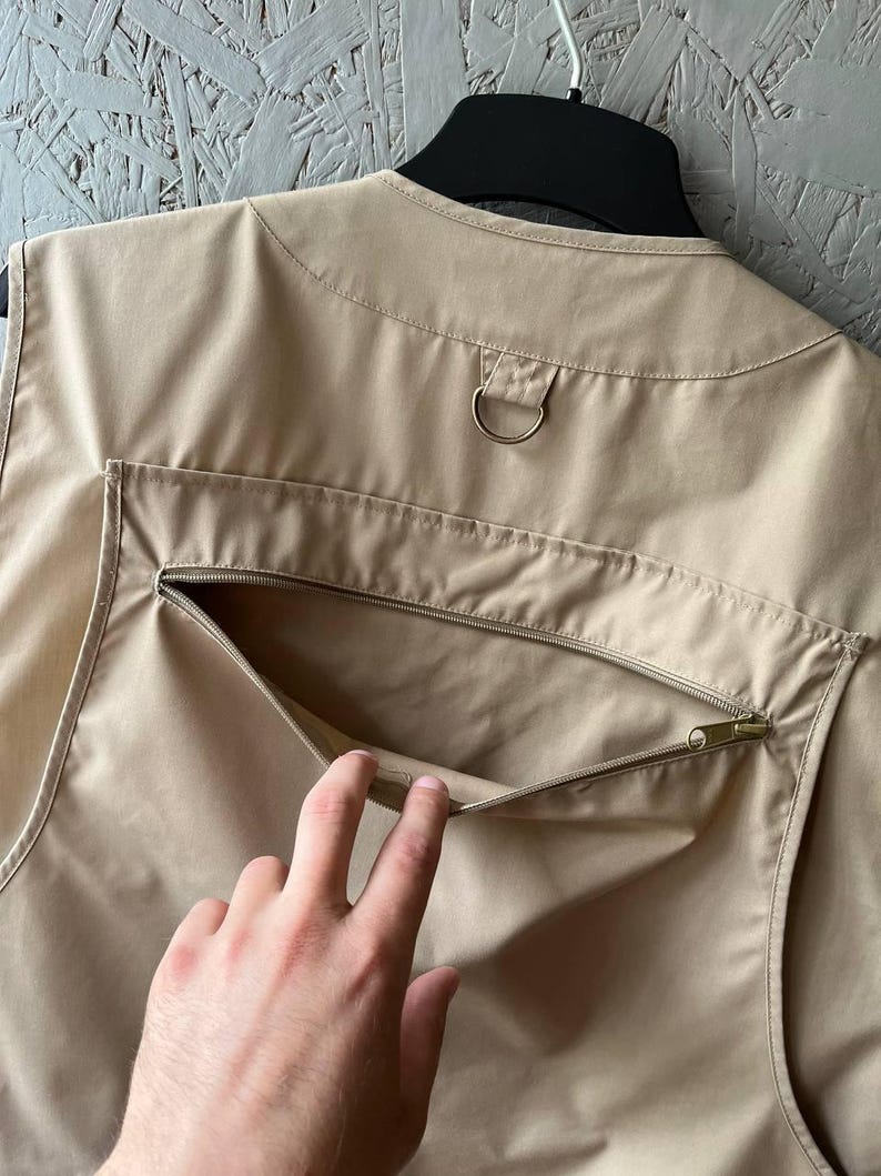 May include: A beige utility vest featuring a sleeveless design with a round neckline. The back of the garment includes a large horizontal zippered pocket and a metal D-ring attachment located just below the collar area for added functionality.