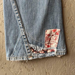 Puede incluir: Vaqueros de mezclilla azul claro con un parche floral. Los vaqueros presentan un dise&ntilde;o de costura &uacute;nico y un dobladillo deshilachado. El parche es rojo y blanco con un estampado floral. Los vaqueros est&aacute;n hechos de mezclilla duradera.