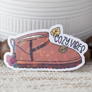 May include: A brown fuzzy boot with a black strap and the text "Cozy Vibes" in pink and black lettering. The boot is surrounded by fall leaves.