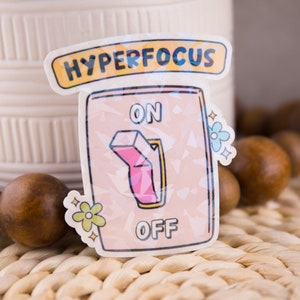 May include: A sticker with the text "HYPERFOCUS" above a switch graphic. The switch is pink and reads "ON" and "OFF". The sticker has a light pink background with floral accents and a clear, iridescent finish.