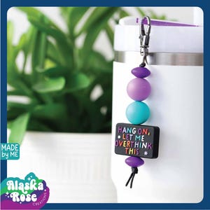 May include: A beaded keychain with a black rectangular charm that reads "HANG ON, * LET ME OVERTHINK THIS *." The keychain features purple, teal, and lavender beads. The background includes a white tumbler and a green plant.