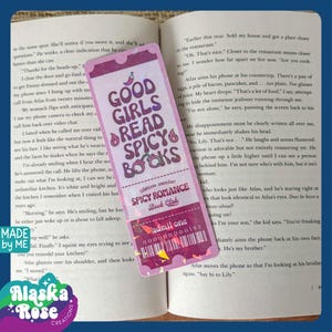 May include: A pink and purple bookmark with the text "Good Girls Read Spicy Books" and a graphic of a red pepper. The bookmark also has the text "Lifetime Member Spicy Romance Book Club" and "Admit One" with a number.
