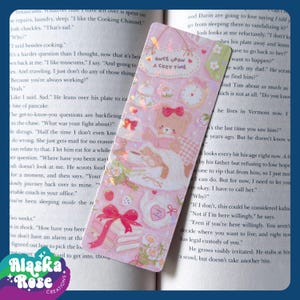 May include: A pink bookmark with a cute bear illustration, bows, and the text "Once Upon a Cozy Time." The design includes a stack of books, a teacup, and a clock. The bookmark is rectangular and has a holographic effect.