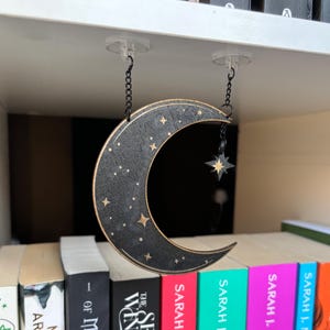 May include: A black crescent moon with gold stars hanging from a shelf with a small gold star charm. The moon is made of wood and has a distressed finish.