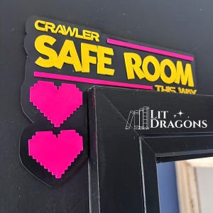 May include: A black rectangular sign with yellow text reading CRAWLER SAFE ROOM THIS WAY and two bright pink pixelated hearts. The sign features a horizontal pink line and is mounted on a black door frame. The text LIT DRAGONS is visible.