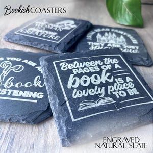 May include: Four black slate coasters with white engraved text. The coasters feature book-themed quotes, including "Between the pages of a book is a lovely place to be." and "If you are a book, I am listening."