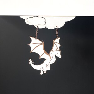 May include: White dragon earrings hanging from a cloud-shaped cutout. The dragon is detailed with wings and a tail, and the earrings are made of metal.