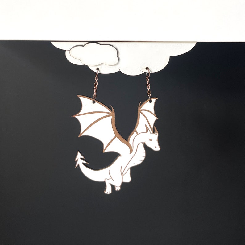 May include: White dragon earrings hanging from a cloud-shaped cutout. The dragon is detailed with wings and a tail, and the earrings are made of metal.