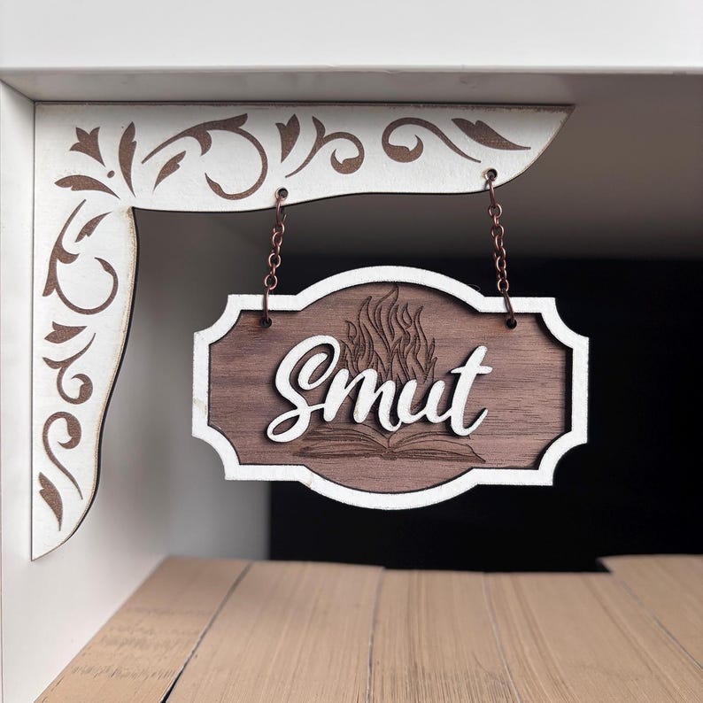 May include: A wooden sign with a white border hangs from a white shelf. The sign has a flame and an open book design with the word "Smut" written in cursive.
