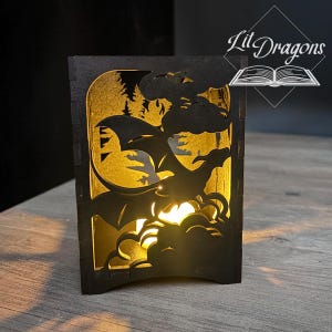 May include: A black wooden lantern with a dragon silhouette design. The lantern features a cutout of a dragon flying through clouds, with a forest scene in the background. The text "Lit Dragons" is visible on the top right.