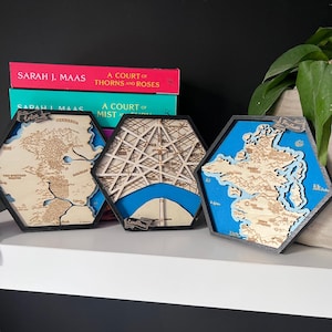 Op de afbeelding: Drie zeshoekige houten kaartlijsten met zwarte randen. De kaarten zijn van fictieve locaties, met blauwe en bruine kleuren. De kaarten zijn getiteld "Enilea", "Lumen" en "Frythuan".
