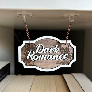 Może przedstawiać: Drewniany znak z białym napisem "Dark Romance" wiszący z sufitu. Znak jest ozdobiony czaszką i wzorami kwiatowymi.