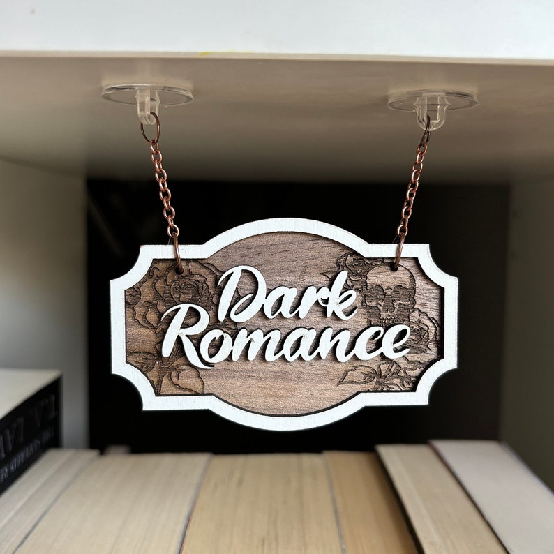 Może przedstawiać: Drewniany znak z białym napisem "Dark Romance" wiszący z sufitu. Znak jest ozdobiony czaszką i wzorami kwiatowymi.