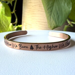 May include: A rose gold cuff bracelet with the engraved text "Hotter We Burn The Higher We Fly" and a flame symbol. The open-ended bracelet is displayed on a white surface, with green foliage in the background.