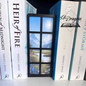 Op de afbeelding: Een zwart houten boekenlegger met een raamontwerp, met een landschap van bergen, een rivier en een heldere zon. De boekenlegger staat tussen boeken met titels als "Heir of Fire" en "Queen of Shadows". Het logo "Lit Dragons" is zichtbaar.