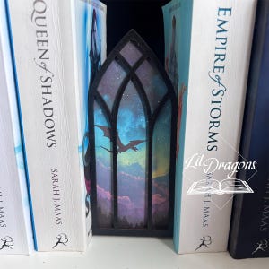 May include: A black wooden bookmark with a stained glass design featuring a dragon flying across a night sky. The bookmark is placed between the pages of books with titles like "Queen of Shadows" and "Empire of Storms". The Lil Dragons logo is visible.