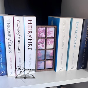 Könnte beinhalten: Ein Buch-Ecke in Fensterform aus Holz mit einer Kirschblüten-Szene, platziert zwischen Büchern. Die Bücher tragen Titel wie "Throne of Glass" und "Heir of Fire" von Sarah J. Maas. Das Logo "Lit Dragons" ist unten zu sehen.