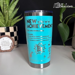 May include: A bright turquoise stainless steel tumbler with a clear lid. The exterior features black text reading NEW ACHIEVEMENT, YOU ARE HYDRATING, REWARD: Dehydration debuff removed. You may continue existing, Crawler, and GOD DAMN IT DONUT.