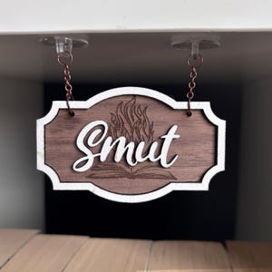 May include: A wooden sign with a white border hangs from two chains. The sign has a brown wood grain finish and features a white cursive word "Smut" with a stylized flame design and an open book underneath.