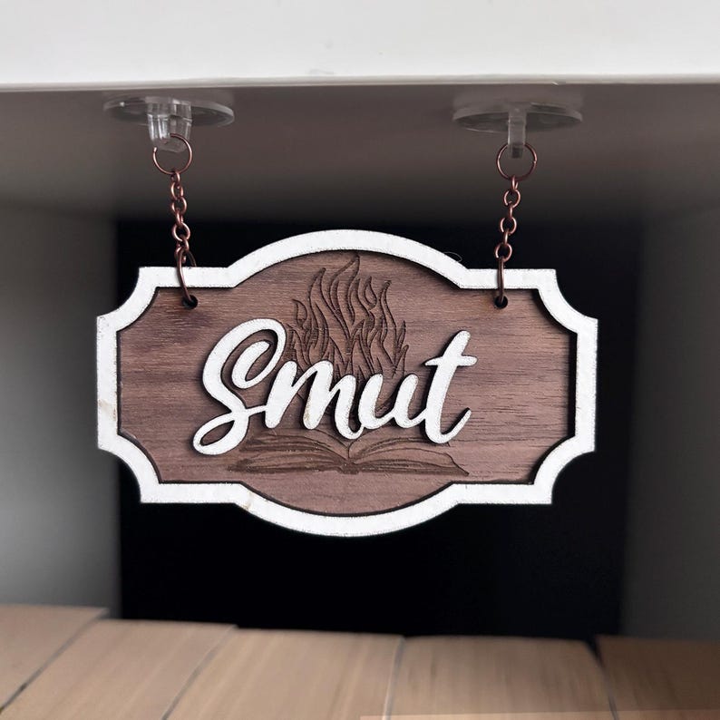 May include: A wooden sign with a white border hangs from two chains. The sign has a brown wood grain finish and features a white cursive word "Smut" with a stylized flame design and an open book underneath.