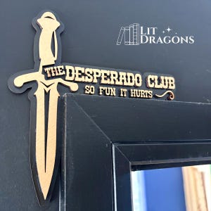 May include: A black and gold decorative door corner accent featuring a vertical sword design. The text reads THE DESPERADO CLUB SO FUN IT HURTS. The piece is mounted on the corner of a black door frame, with a small logo reading LIT DRAGONS.