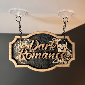 Może przedstawiać: Drewniany znak z napisem "Dark Romance" w ozdobnym kroju pisma. Znak jest czarny z fakturą drewna i przedstawia czaszkę i r&oacute;że.