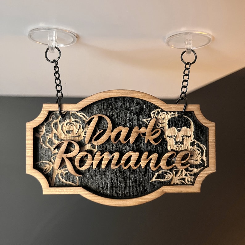 Może przedstawiać: Drewniany znak z napisem "Dark Romance" w ozdobnym kroju pisma. Znak jest czarny z fakturą drewna i przedstawia czaszkę i r&oacute;że.