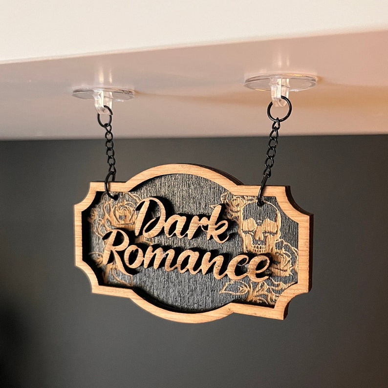 Może przedstawiać: Drewniany znak z napisem "Dark Romance" w ozdobnym kroju pisma. Znak jest zawieszony na suficie za pomocą dw&oacute;ch przezroczystych przyssawek i czarnych łańcuch&oacute;w. Znak ma ciemne drewniane tło z kwiatowym wzorem i motywem czaszki.
