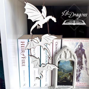 May include: Three wooden dragon ornaments suspended by a chain, with the text "Lit Dragons" in the upper right corner. The dragons are positioned in front of a bookshelf with books, including "Heir of Fire" and "Crown of Shadows".