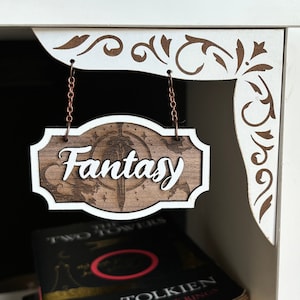 May include: A wooden sign with white trim hangs from two chains. The sign says "Fantasy" in white cursive letters on a brown background with a stylized sword and stars.