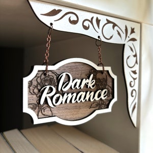 Może przedstawiać: Drewniany znak z białą ramką wisi na dw&oacute;ch miedzianych łańcuchach. Znak jest ozdobiony wzorem kwiatowym i napisami "Dark Romance" w białym, ozdobnym piśmie.