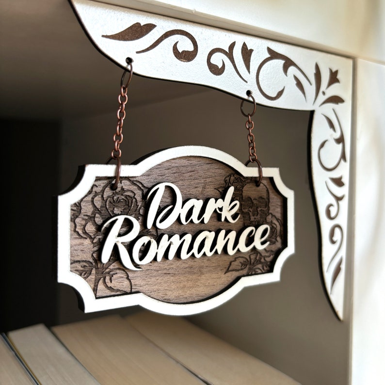 Może przedstawiać: Drewniany znak z białą ramką wisi na dw&oacute;ch miedzianych łańcuchach. Znak jest ozdobiony wzorem kwiatowym i napisami "Dark Romance" w białym, ozdobnym piśmie.